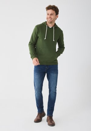 Sweat à capuche vert avec une poche kangourou, cordons blancs, porté sur une chemise blanche. Associé à un jean bleu et des bottes marron.