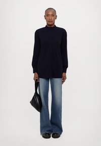 Marineblå bluse med lange ærmer og høj krave, parret med lys blå flared jeans og sorte platform sko, mens hun holder en sort handbag.