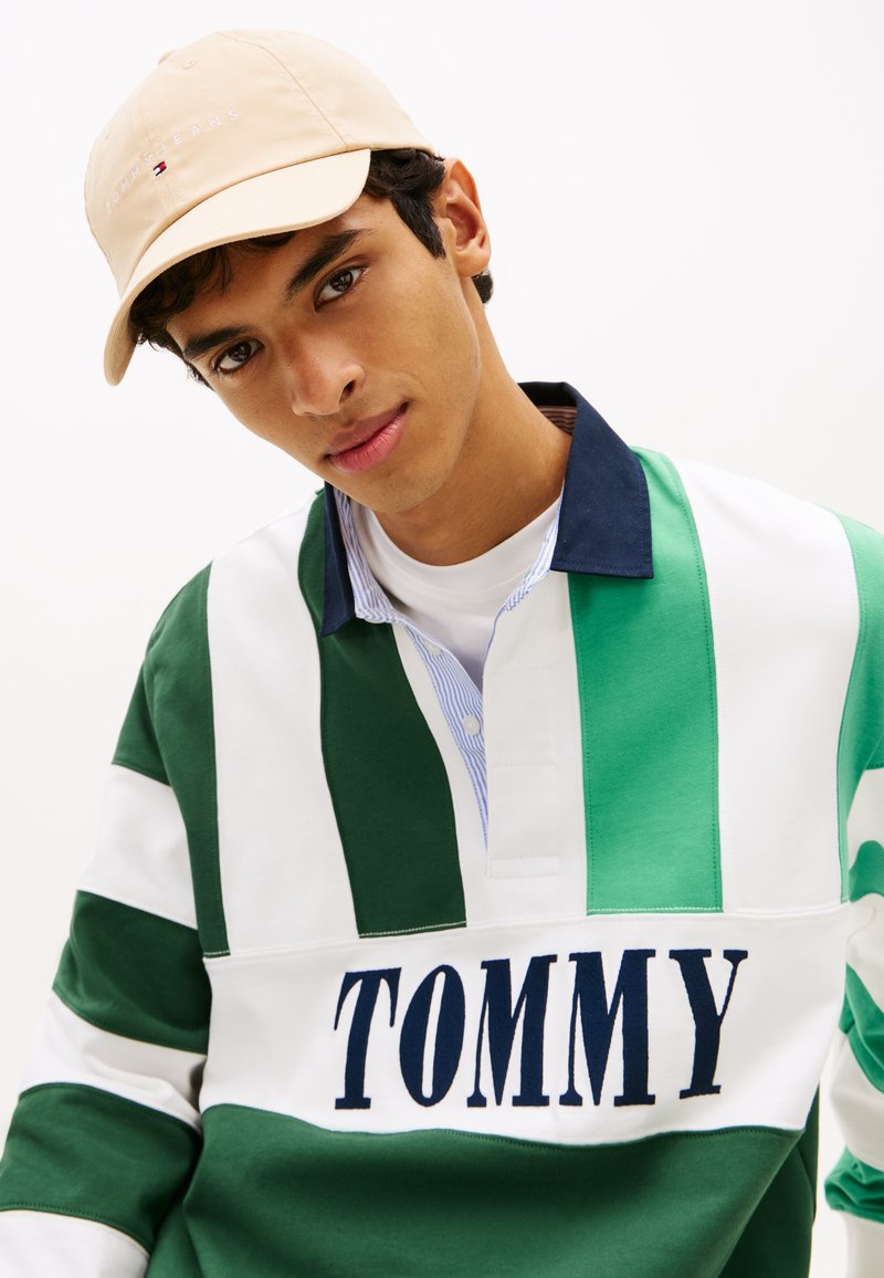 Ρουαγιάλ ριγέ μπλούζα ράγκμπι σε πράσινο και λευκό με μπλε γιακά, διακοσμημένη με τη λέξη "TOMMY" σε μπλε γράμματα· συνδυασμένη με μπεζ καπέλο μπέιζμπολ.