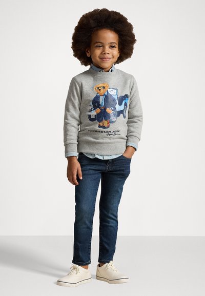 Sudadera gris con un gráfico de un oso con chaqueta azul, jeans azules y zapatos beige. El diseño incluye colores vibrantes y detalles elaborados.
