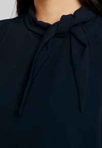 Marinblus i ett mjukt, texturerat tyg, med en knutdetalj vid halsen och en hög krage. Slät fall och figursydd design.