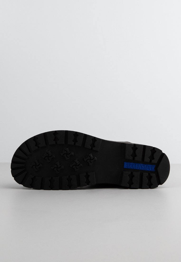 Schwarze Schuhsohle mit tiefen Profilrillen und einem blauen Birkenstock-Logo auf der Ferse, vor einem schlichten grauen Hintergrund gezeigt.