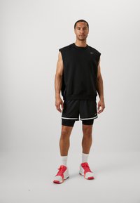 Black sleeveless sweatshirt and black shorts with white trim. Pink and white athletic shoes. Model stands against a plain background.