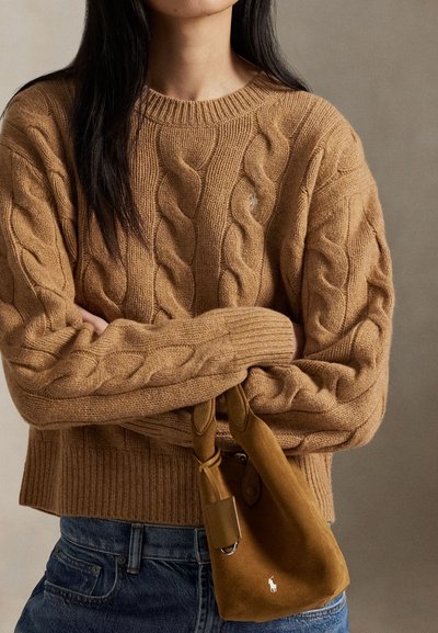 Žena nosící béžový svetr s pleteným vzorem, držící malou hnědou semišovou tašku s bílým logem, v kombinaci s modrými džínami, na neutrálním pozadí.