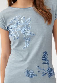 Jasnoniebieska bluzka z krótkim rękawem z aplikacjami kwiatowymi z cekinów w różnych odcieniach niebieskiego na przodzie. Miękka, gładka faktura.
