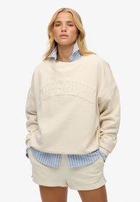 Sweatshirt crème avec un logo "SUPERDRY" embossé, superposé sur une chemise col bleu à rayures. Assorti à un short clair. Texture de tissu douce.