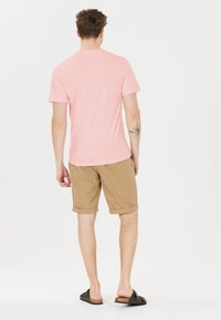 Vaaleanpunainen lyhythihainen t-paita, vaaleat rullatut shortsit, mustat sandaalit. Malli näkyy takaa, vasemmassa kädessä näkyy tatuointi. Suora kangas.