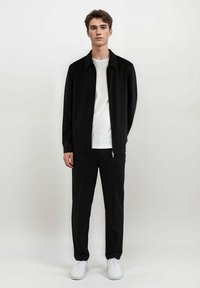 Svart jakke med glidelås, hvit t-skjorte under, matchende svarte bukser og hvite joggesko. Jevn tekstur, rene linjer, minimal design.