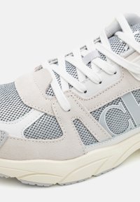 Sneaker a rete grigio chiaro con accenti in suede beige, lacci bianchi e suola testurizzata. Presenta un dettaglio del logo sul lato e un colletto imbottito.