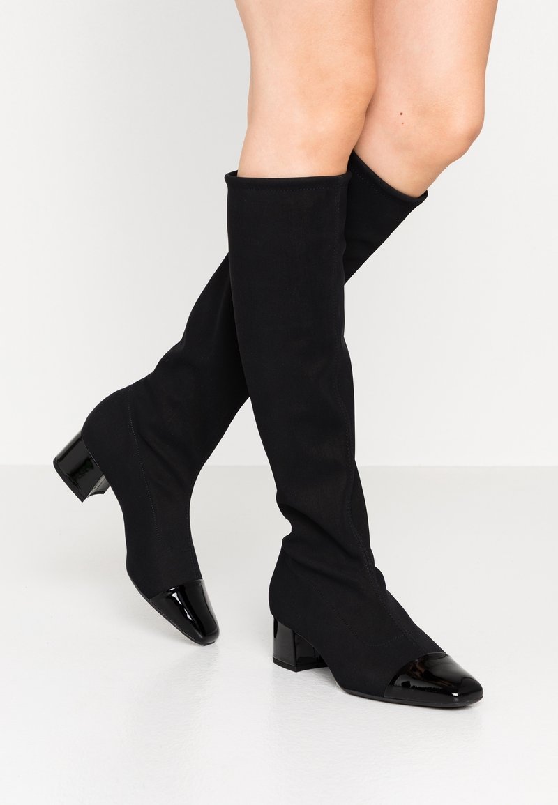 Jambes portant des bottes noires montantes à talons blocs avec des bouts brillants noirs, debout sur un sol blanc devant un fond blanc uni.