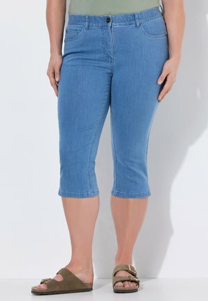 Vrouw draagt lichtblauwe cropped spijkerbroek en olijfgroene sandalen met gesp, staand tegen een witte muur.