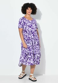 Lila Batik-Kleid aus Baumwolle, mit kurzen Ärmeln, Taschen und einem gestuften Design für eine entspannte Passform. Kombiniert mit weißen Riemchensandalen.