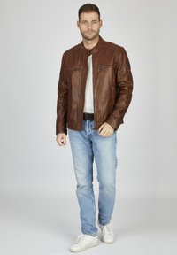 Giacca di pelle marrone con zip frontale, due tasche sul petto e dettagli in rilievo sulle spalle, abbinata a jeans azzurri chiari e sneakers bianche.
