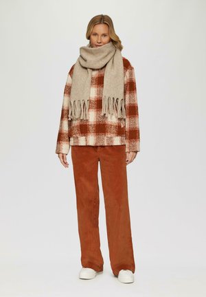 Oversized Hemd in braun-weißem Karomuster mit langen Ärmeln, kombiniert mit rostfarbenen Cordhose und einem beigen, fransigen Schal. Weiße Sneaker.