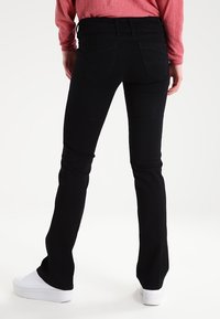 Pantalones vaqueros negros con diseño de pierna acampanada, hechos de denim. Presentan bolsillos traseros y una cintura con passes para cinturón. Combinados con zapatos blancos.