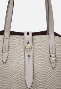 Sac fourre-tout en cuir beige texturé avec deux longues poignées, ferrures décoratives en or, et une sangle centrale ornée de découpes.