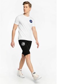 Weißes Baumwoll-T-Shirt mit NASA-Logo, kombiniert mit schwarzen Shorts mit einem Logo, ergänzt durch hochhackige weiße Sneakers mit roten Akzenten.