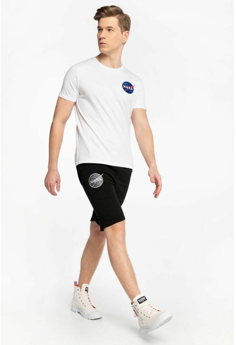 Weißes Baumwoll-T-Shirt mit NASA-Logo, kombiniert mit schwarzen Shorts mit einem Logo, ergänzt durch hochhackige weiße Sneakers mit roten Akzenten.