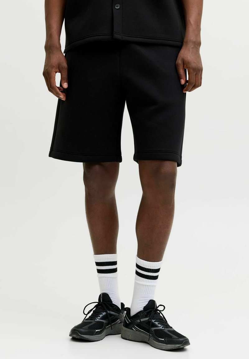 Modèle masculin portant un short décontracté noir, des chaussettes blanches à rayures noires et des baskets noires, debout sur un fond blanc.