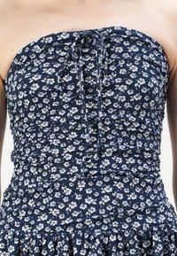 Jūrinės gėlės rašto suknelė tamsiai mėlyna, su mažomis baltomis gėlėmis, be petnešėlių, turinti susiraukšlėjimą, su raišteliu iškirptėje ir pamušta kraštu.