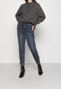 Frau in einem dunkelgrauen Strick-Hoodie, hochgeschnittenen blauen Jeans mit einem schwarzen Gürtel und schwarzen, hochhackigen Stiefeletten, die vor einem schlichten Hintergrund steht.