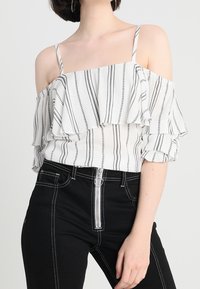 Top bianco scollato con righe verticali nere, caratterizzato da volant a strati, spalline sottili e maniche a sbuffo. Abbinato a jeans neri a vita alta.