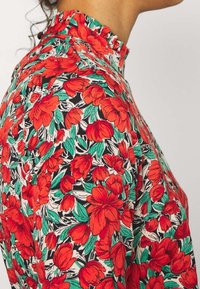 Blumenbluse mit einem mehrfarbigen Muster aus roten Blumen und grünen Blättern auf schwarzem Hintergrund, mit hohem Rüschenausschnitt und weicher Textur.