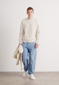 Cremfarbener, gerippter Pullover, hellblaue Jeans mit lockerer Passform, weiße Sneakers, hält eine hellbeige Jacke. Schlichte, neutrale Farbpalette.