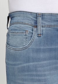 Los jeans de mezclilla azul claro presentan un bolsillo trasero, costuras en contraste y una cinturilla de altura media. La tela muestra una textura cepillada.