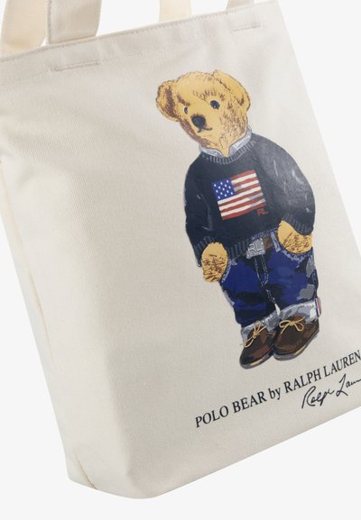 Platnena torba sa ilustracijom medvjeda u tamnoplavom puloveru s američkom zastavom, plavim trapericama i smeškim cipelama. Ispod se nalazi natpis "POLO MEDVJED od RALPH LAUREN-a."