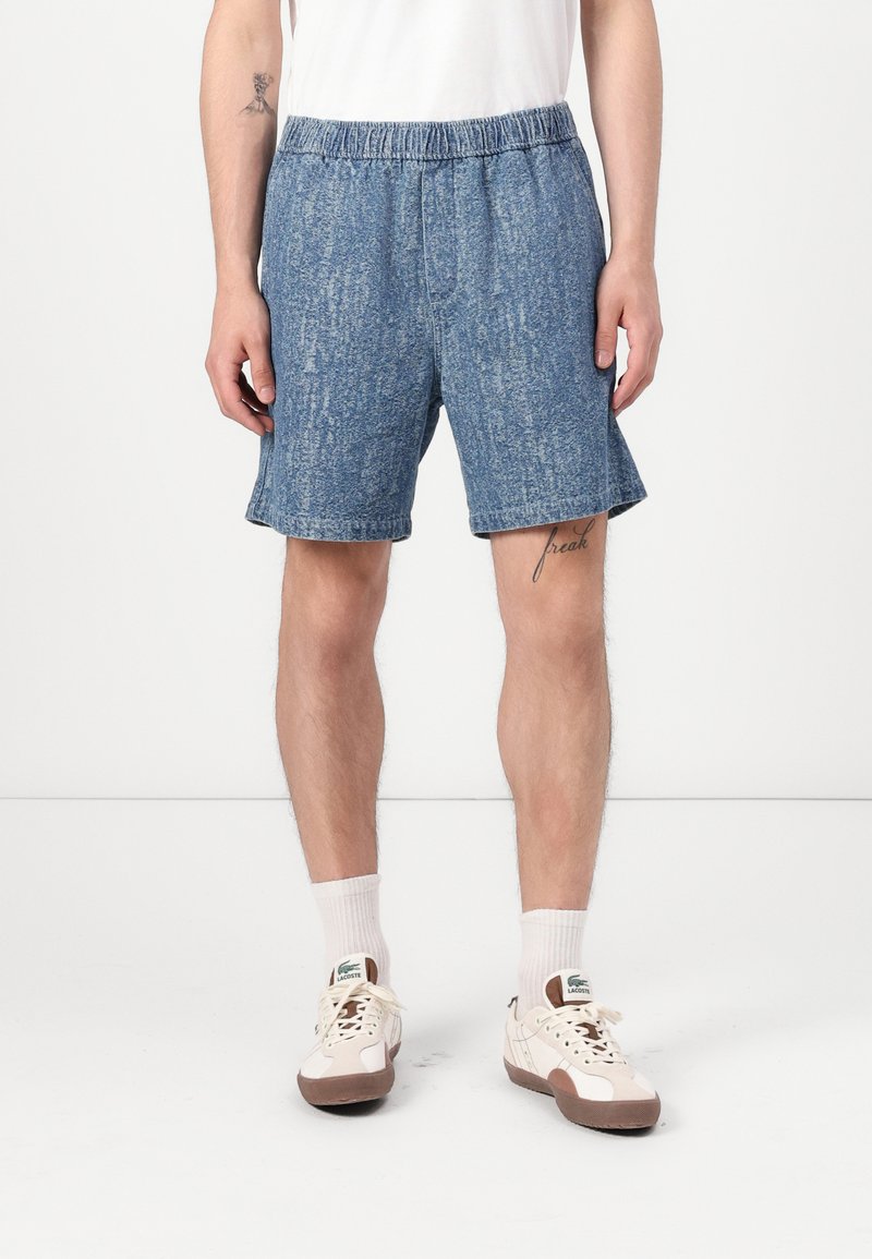 Bermudas de mezclilla en azul claro con una textura sutil, que cuentan con una cintura elástica y bolsillos laterales, combinadas con calcetines deportivos blancos y zapatillas.