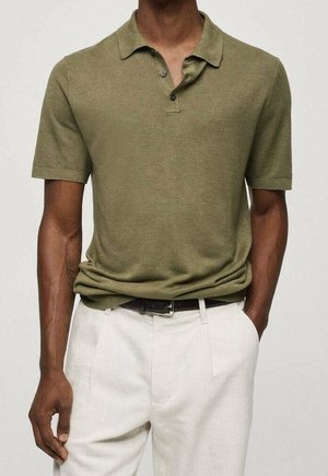 Uomo con polo verde oliva a maniche corte infilata in pantaloni bianchi a vita alta con cintura nera, in piedi con una mano in tasca.