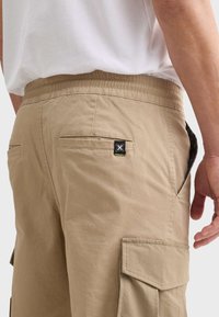 Khaki cargo šortky s elastickým pasem, dvěma postranními kapsami a štítkem s černými a zelenými akcenty na zadní kapse.