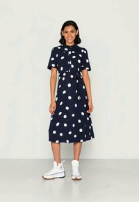 Γυναίκα με χαμόγελο, ντυμένη με navy blue midi φόρεμα με μεγάλα λευκά πουά και λευκά sneakers ψηλής ankle, στέκεται μπροστά σε απλό φόντο.