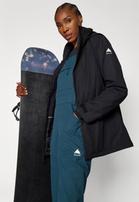 Czarna kurtka snowboardowa z kapturem, zestawiona z turkusowymi ogrodniczkami. Model trzyma czarny snowboard z niebiesko-fioletowym wzorem teksturowym.