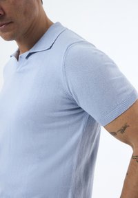 Polo de punto azul claro con mangas cortas. Presenta un cuello clásico y una textura suave, resaltando una silueta ajustada.