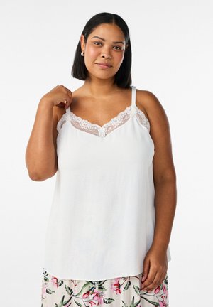 Žena so stredným odtieňom pokožky a čiernymi vlasmi, oblečená v bielom saténovom topánke s čipkovým lemom a nohaviciach s kvetinovým vzorom, stojaca na bielom pozadí.