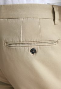 Beige chinos with a smooth, woven texture. Features a back pocket with a black button and subtle stitching details above the pocket.