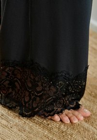 Fekete ruhadarab csipkés szegéllyel az alján, bonyolult virágmintákkal és sima, puha textúrával. Lábujjak láthatók egy mintás padlón.