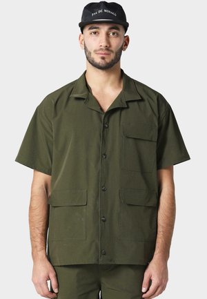 Forest green short-sleeve shirt, button-down front, two front pockets, smooth fabric, relaxed fit, worn with matching pants and a black cap.