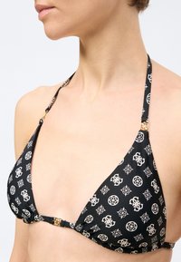 Γυναίκα που φοράει μαύρο τρίγωνο μπικίνι τοπ με λευκά γεωμετρικά και λογότυπα σχέδια και μεταλλικά κούμπωμα χρυσού τόνου στον λαιμό και μπροστά.