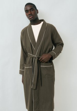 Joven de piel oscura con una túnica verde oliva con ribetes blancos, ajustándose el cinturón, de pie frente a un fondo claro y sencillo.