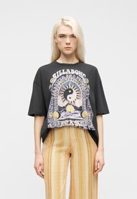 Ξανθιά γυναίκα που φοράει φαρδιά μαύρη μπλούζα Billabong με γραφικό ήλιου γιν-γιανγκ και ριγωτό μπεζ παντελόνι, στέκεται μπροστά σε λευκό φόντο.