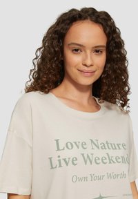 Cremefarbenes, oversized T-Shirt mit Rundhalsausschnitt und kurzen Ärmeln, mit grüner Schrift: "Liebe die Natur, lebe das Wochenende, erkenne deinen Wert."