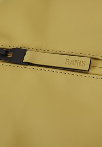 Sárga esőkabát sima, matt felülettel. Kontrasztos fekete cipzárral és a "RAINS" szóval domborítva ellátott márkás húzókával rendelkezik.