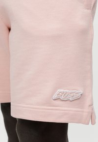 Ανοιχτό ροζ σορτς με μικρό λευκό κεντητό λογότυπο "BOSS" κοντά στο τελείωμα, φορεμένο σε σκούρο μανεκέν πόδι.