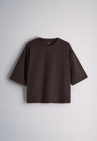 Mörkbrun T-shirt med kort ärm och rund halsringning, lös passform, hängande mot en enfärgad ljusgrå bakgrund.