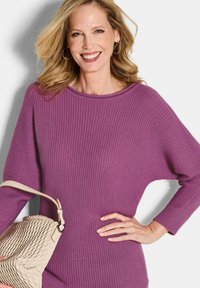Lächelnde Frau mit welligem blonden Haar trägt einen lila gerippten Pullover und hält mit einer Hand eine beigefarbene strukturierte Handtasche, während sie mit der anderen Hand auf ihrer Hüfte steht.