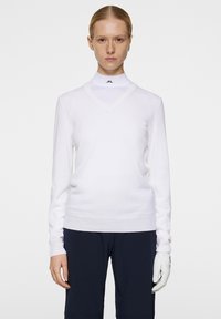 Valkoinen v-kaula-aukko kietaisu neulepaita pitkillä hihoilla, jossa on tiukka istuvuus ja ribloomit hihansuut. Yhdistetty turtleneck-paidan kanssa ja tummien housujen kanssa.
