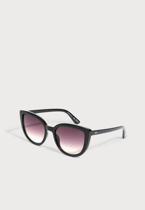 Schwarze Cat-Eye-Sonnenbrille mit verlaufend violett getönten Gläsern, die auf einer hellgrauen Oberfläche liegt.